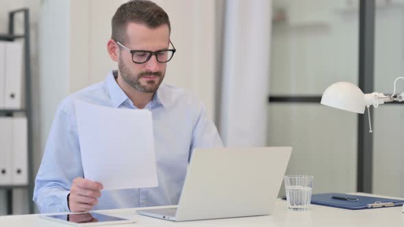 Middle Aged Man Reading Documents While Working on Laptop alt