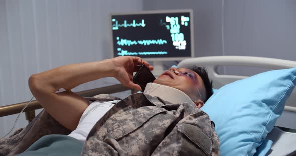 Injured Asian Soldier with Neck Collar Lying in Hospital Bed and Talking on Smartphone alt
