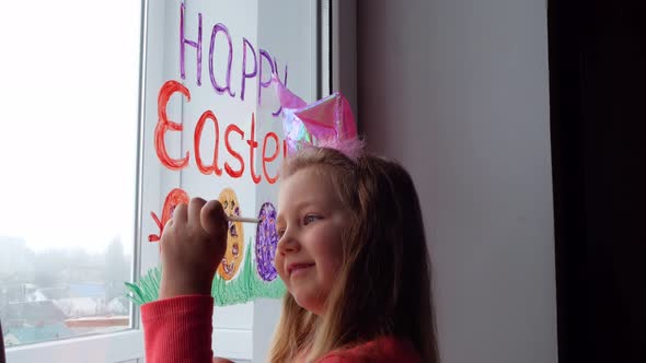 Cute little girl in bunny ears painting Easter eggs with green spring grass on window at home alt