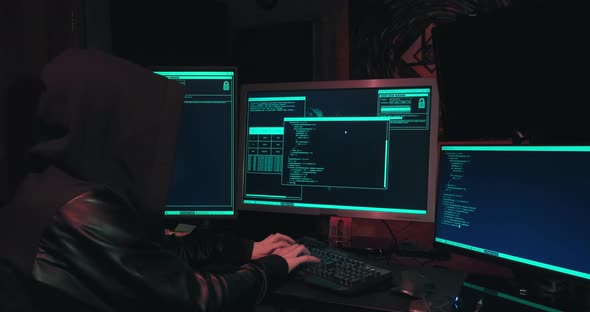 Hacker in a Hood Sits in Front of Computer Screens and Hacks Databases alt