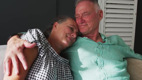Caucasian senior couple hugging each other while sitting on the couch at home alt