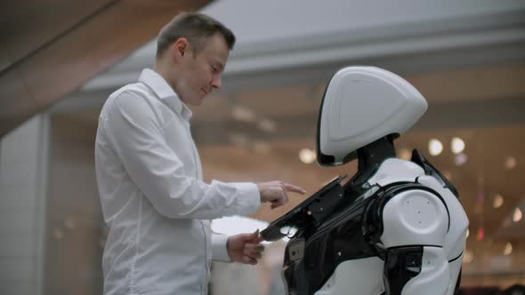 A Man Stands with a Robot Bot and Asks Him Questions and Asks for Help By Clicking on the Screen on alt