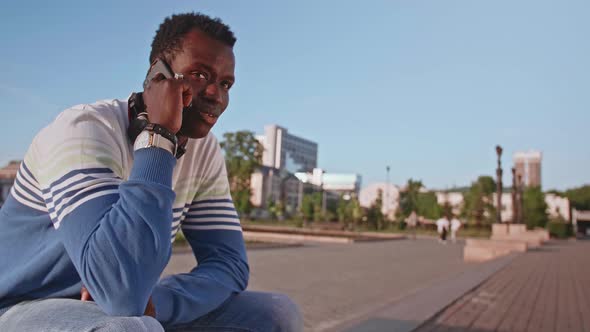 A Young Black Student in Stylish Clothes Sits and Talks on a Smartphone in the City