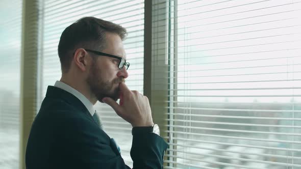 Focused Creative Man in a Suit Stands at the Window in the Office and Reflects the Thinking Process alt