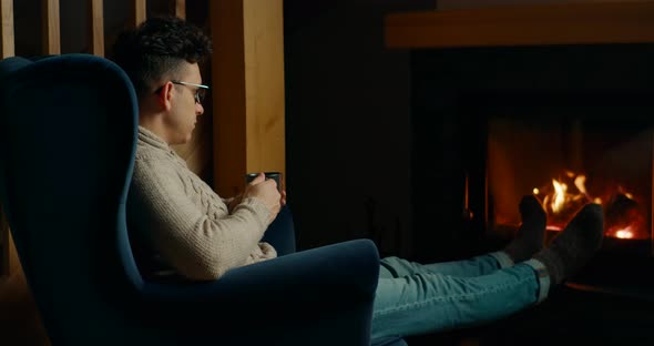 Relaxed Man Warms at Fireplace Sitting in Armchair and Drinking Hot Tea at Night