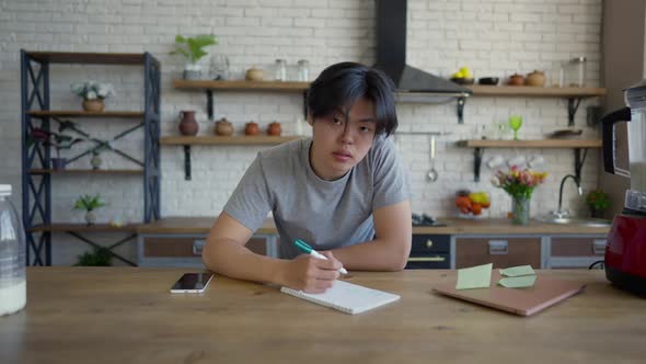 Wide Shot of Young Man Writing in Slow Motion and Looking at Camera Sitting at Kitchen Table alt