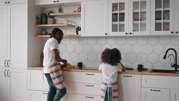 African American Man Wearing Apron Cutting Oranges at Kitchen His Little Daughter Helping Him with alt