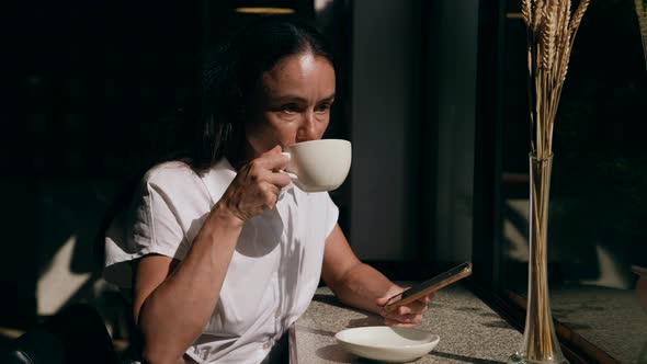 Middle Aged Woman is Enjoying Cup of Coffee in Cafe at Sunny Day and Surfing Internet By Cellphone alt