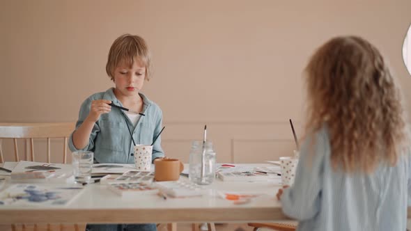 Group of Kids and Their Teacher Leaning Over Table with Watercolor Paints and Painting Selfmade Clay alt