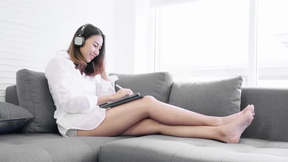 Young Asian woman using digital tablet and headphone on sofa at home living room. Work at home