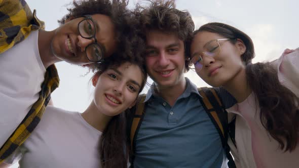 Low Angle View of Happy Diverse Friends Hug and Look Down at Camera Outdoors alt