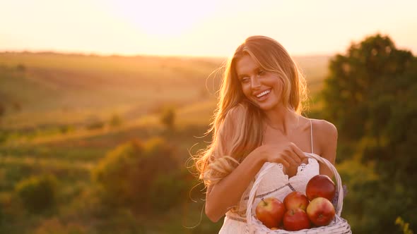 Beautiful sexy blonde girl in white dress posing in a field at sunset with a basket of fruit	 alt