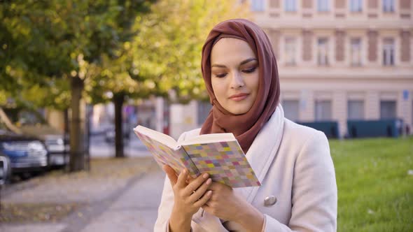 A Young Beautiful Muslim Woman Reads a Book with a Smile in a Street in an Urban Area  Closeup alt