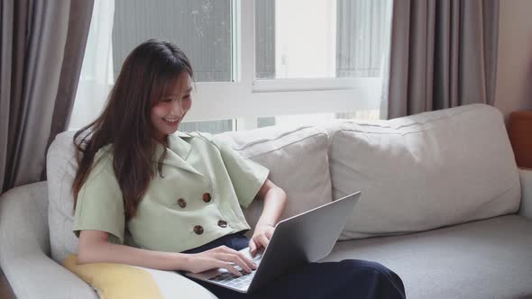 Happy young asian woman sitting on sofa using laptop to video call or facetime to talk with friend alt