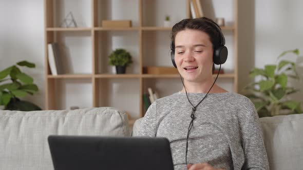 Attractive Woman Resting on Couch Wear Headphones Looks at Laptop Screen Greeting Friend or Family alt