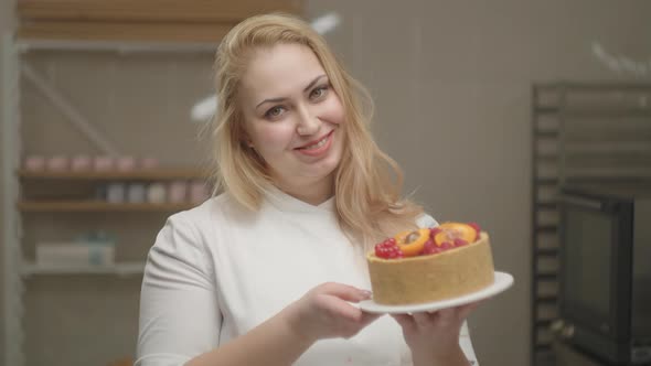 Smiling Confectioner Woman in White Coat Holding Cheesecake Decorated with Apricot and Berries