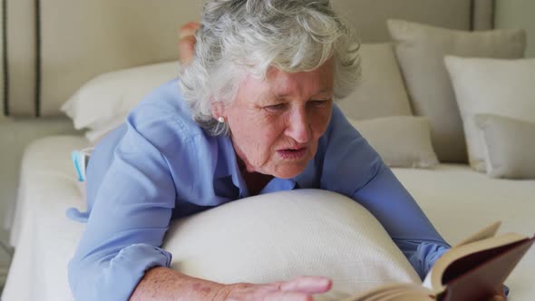 Happy caucasian senior woman at home enjoying reading book, lying on bed in sunny bedroom alt
