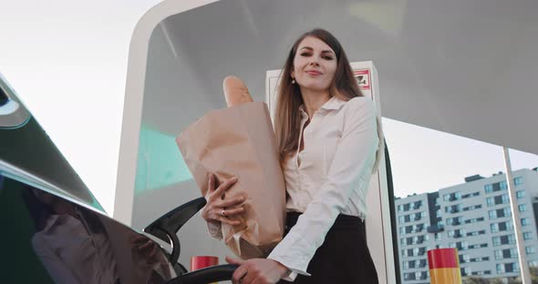 A Woman Stands With A Package Of Products Near Her Electric Car And Waits For alt