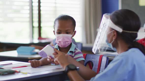 Diverse medical worker showing schoolgirl how to measure temperature, all wearing face masks, alt