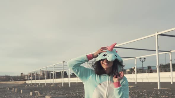 Cheerful Girl Is Wearing Blue Kigurumi of Unicorn and Jumping on Beach alt