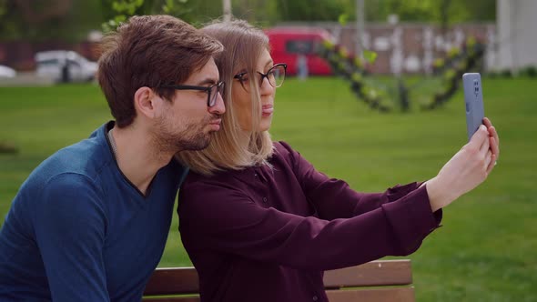 Two friends with glasses sit on the bench outside take selfie alt