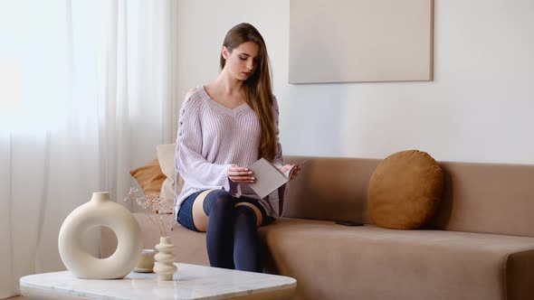 Young Woman At Home Sitting On Sofa In Living Room And Reading