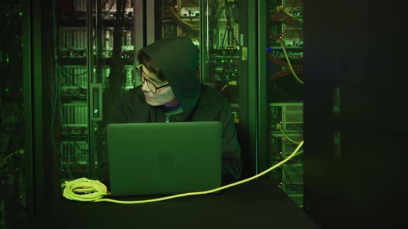 Asian male hacker in hoodie using laptop by computer servers alt