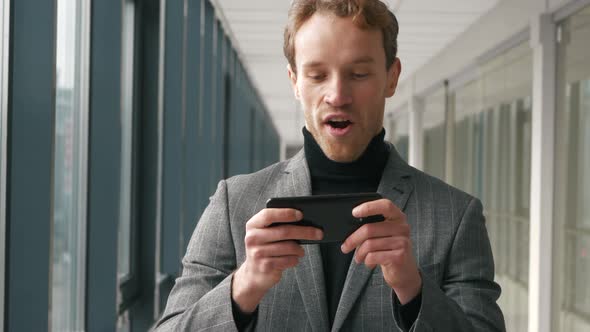 Close up bussisman is enjoying playing video game in smartphone standidng in office. Young man playi alt