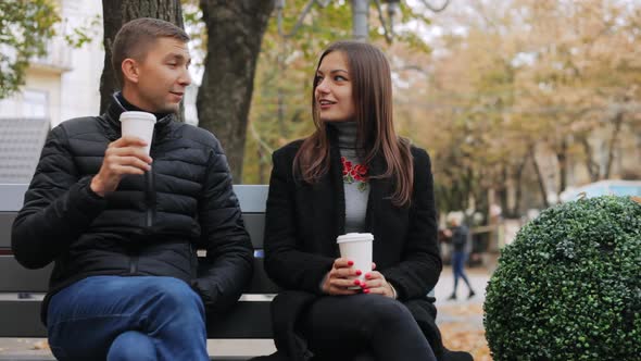Happy Couple with Paper Glasses in Hands Talking on City Bench on the Street alt