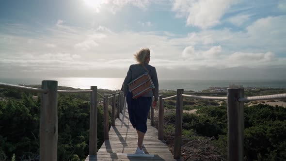 A Middleaged European Woman Artist with an Easel on Her Shoulder Walks Towards the Ocean alt