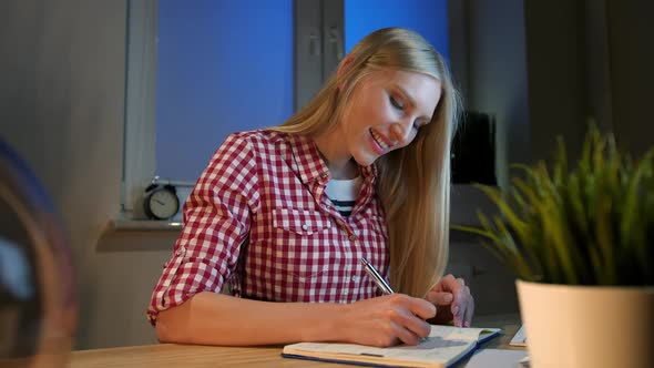 Cheerful Female Writing in Daily Planner. Lively Young Blond Woman in Casual Checkered Shirt Sitting alt