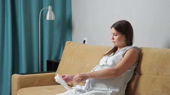 Pregnant Woman with Big Belly Prepares Baby Clothes While Sitting on Sofa alt