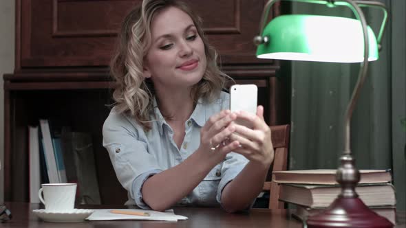 Relaxed Youn Woman Sitting at the Table with Books and Taking Funny Selfies with Her Phone
