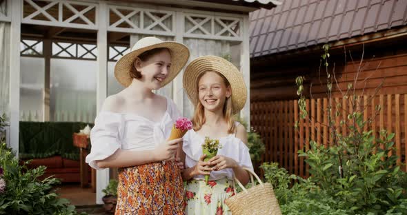 Two Cheerful Teen Girls in Straw Hats and Rural Dresses with Flowers in Their Hands Laughing and alt