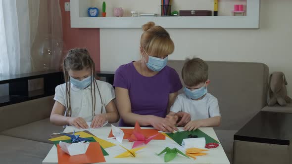 Mother, Son and Daughter in Medical Masks Make Crafts From Colored Paper in the Room. Social
