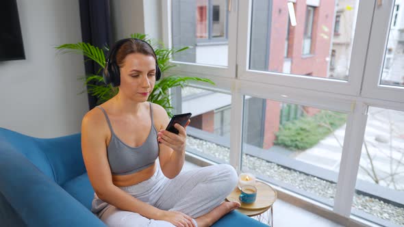 Woman in Wireless Headphones Listening to Music and Using Mobile Apps on Smartphone Sitting on Sofa alt