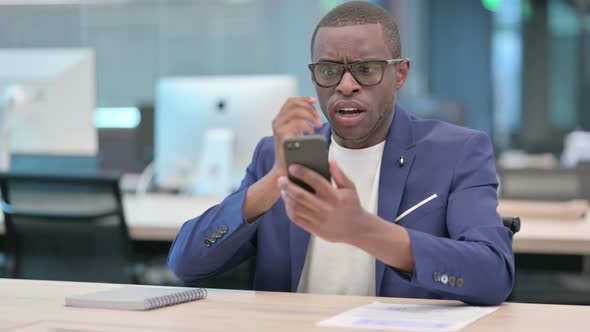 African Businessman Reacting to Loss on Smartphone in Office alt