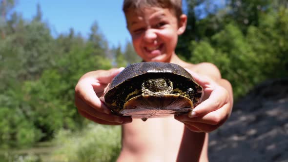 Boy Holds Turtle in Arms and Smiles Viciously on River with Green Vegetation alt