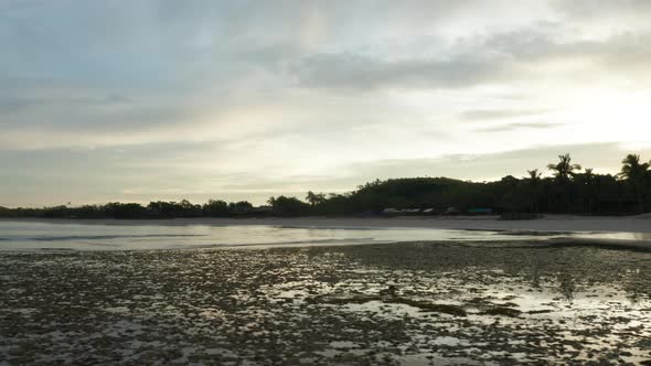 Low tide in tropical bay with silhouette shore from rising sun behind it, drone alt