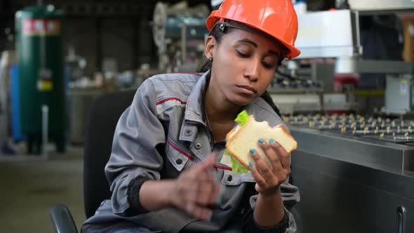 A young black woman in an orange helmet eats a sandwich in a production facility alt