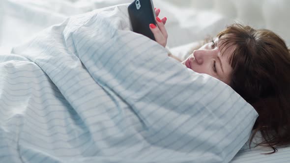 Sleepy Brunette Woman Lies on the Bed and Uses the Phone in the Morning Read Some Information