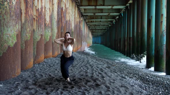 a Barefoot Woman Dances Under the Pillars of the Bridge Against the Background of the Incoming Waves alt
