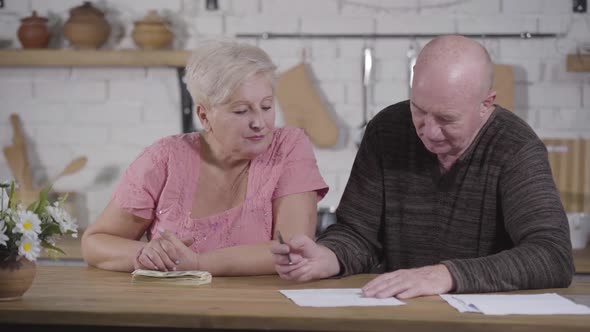 Portrait of Senior Caucasian Woman Looking at Her Husband Writing Down Budget. Couple Settling alt