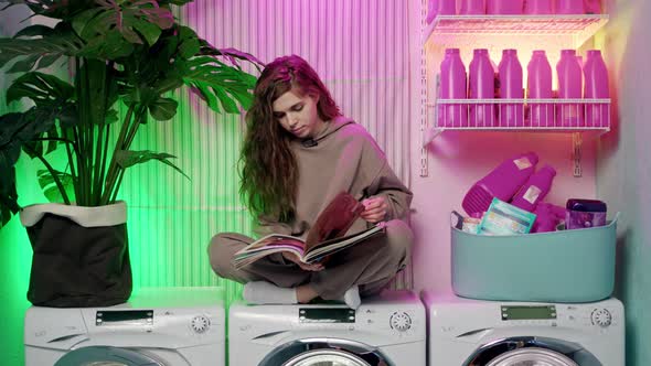 Beautiful Young Woman with Long Brown Wavy Hair in Beige Tracksuit Sits on Washing Machines and alt