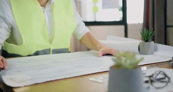 Architect in Helmet and Vest Sitting at the Desk in Workroom and Browsing Ready Blueprint alt