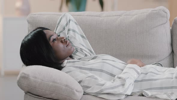 Young Pensive Calm Dreaming African American Woman Resting on Comfortable Sofa in Living Room alt