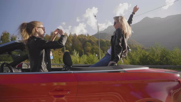 Two Caucasian Girls in a Cabriolet are Photographed on a Smartphone and Smiling