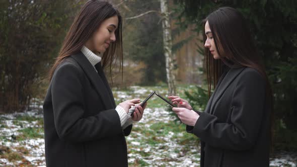 Two Beautiful caucasian twins women in winter wood outdoors using smartphone or cell phone. Tapping,
