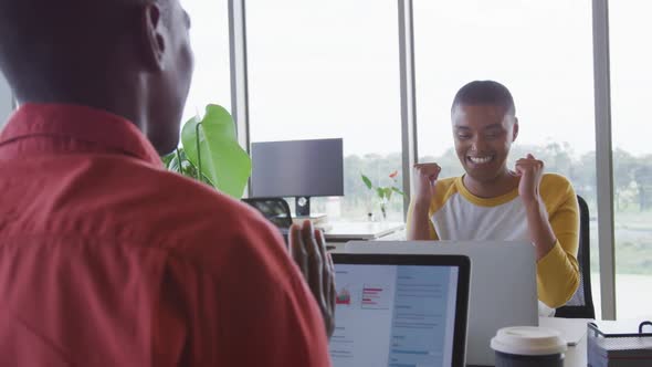 Happy african american creative businesswoman celebrating at desk in office with male colleague alt