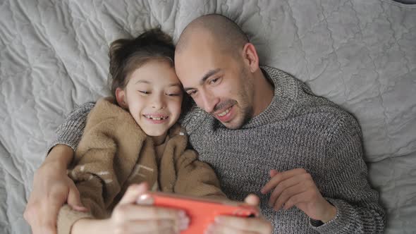 Father and Daughter Lie on the Bed and Communicate Via Video Link on a Smartphone alt